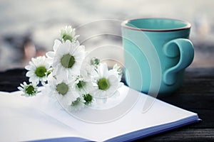 Coffee cup or tea cup and white daisy flowers with blank paper book on bright background. Notebook, flower and morning coffee.