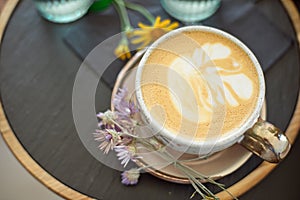 Coffee cup with white gold stands on a saucer with a black tray of flowers. Flat lay.