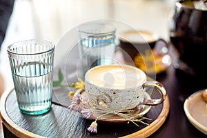 Coffee cup with white gold stands on a saucer with a black tray of flowers. Flat lay.