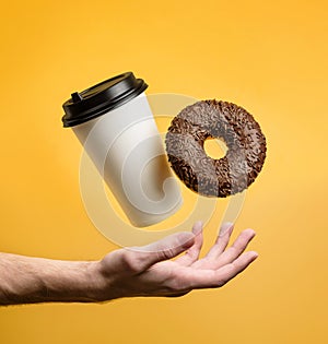 A coffee glass and a chocolate donut levitate above an outstretched palm on a yellow background. Takeaway food concept