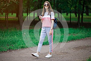 Coffee on go. Beautiful young woman in sunglasses holding coffee cup and smiling while walking