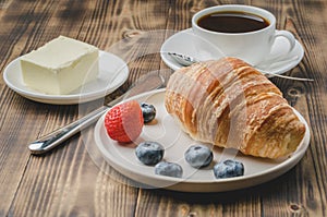 Coffee hous cup, croissant with berries in white bowl and butter on wooden background. Healthy breakfast with fresh berries