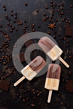 Coffee ice cream on a stick, chocolate slices, scattered coffee beans, dark brown background. Top view, space for text.