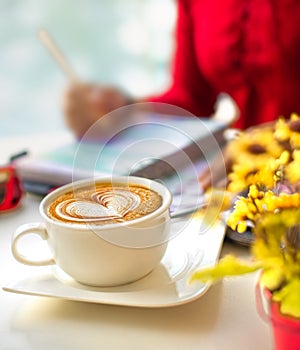Coffee Latte Art with Blur Yellow Flower in Front and Woman Wearing Red Dress Writing on Book. Selective Focus.