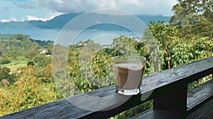 Coffee latte glass on rail above green valley in the mountains. Good morning. Caffeine drink in Kintamani Bali.