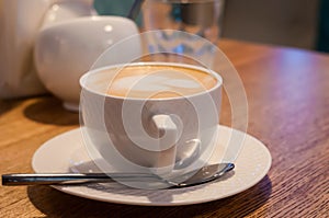 Coffee with milk foam in white cup with sauser and spoon on wooden table in cafe