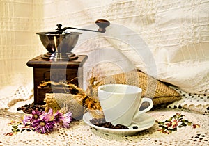 Coffee mill with burlap sack of roasted beans and white cup of coffee on the white knitted linen table-cloth