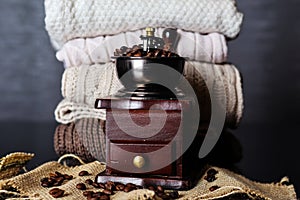 Coffee mill with roasted black coffee beans on desk at home and sweaters in background