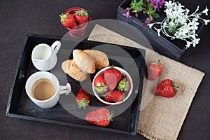 Coffee, mini French pastries and strawberries on wooden tray over black table. White and purple flowers in a decorative wooden