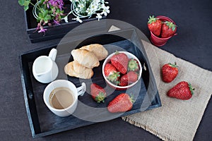 Coffee, mini French pastries and strawberries on wooden tray over black table. White and purple flowers in a decorative wooden