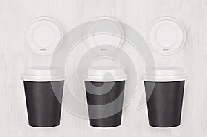Coffee mockup - set of three black paper cups and blank white caps on white wood board, top view.