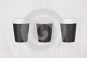 Coffee mockup - set of three blank black paper cups with white caps on white wood board, top view.