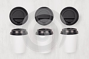 Coffee mockup - set of three white paper cups and blank black caps on white wood board, top view.