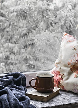 Coffee mug, book, pillows and a plaid on the light wooden surface against window with rainy day view. Vintage style.