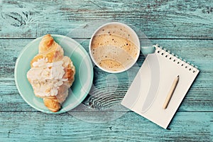 Coffee mug with croissant and empty notebook and pencil for business plan and design ideas on turquoise rustic table from above photo