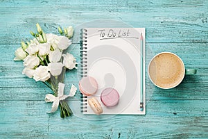 Coffee mug with macaron, white flowers and notebook with to do list on blue rustic table from above. Beautiful breakfast. Flat lay