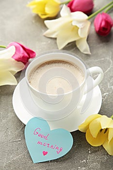 Coffee mug with tulip flowers and notes good morning on grey background, breakfast on Mothers day or Womens day. Vertical photo