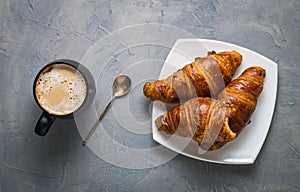 Coffee and pastries: a black mug with coffee, a silver spoon and a croissant on a saucer on a gray background.