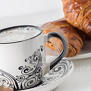 Coffee and pastries: a white coffee mug with a pattern, a silver spoon, fresh croissants on a saucer.