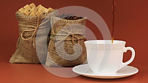 Coffee is poured into a cup and behind stands coffee beans in burlap bag and next sugar in burlap bag on brown