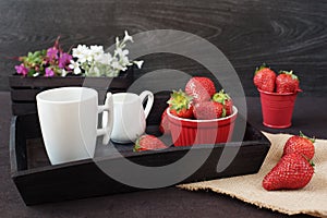 Coffee and strawberries on wooden tray over black table. White and purple flowers in a decorative wooden crate. Black background