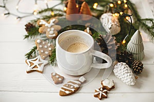 Coffee in stylish cup with gingerbread cookies, pine cones and warm lights on white wooden table