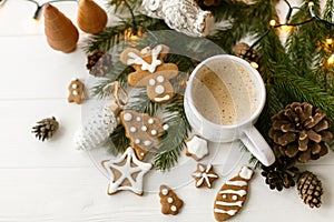 Coffee in stylish cup with gingerbread cookies, pine cones and warm lights on white wooden table