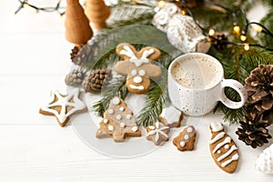 Coffee in stylish cup with gingerbread cookies, pine cones and warm lights on white wooden table