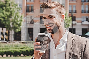Coffee time. Portrait of confident brown-haired man holding coffee cup and smiling aside while walking outdoors