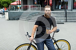 Coffee to go. Side view of young bearded man drinking coffee while sitting on his bicycle outdoors