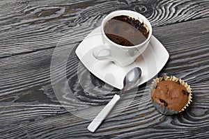 Coffee in a white cup stands on a saucer. Nearby lies a muffin, and a spoon on a saucer. On brushed pine boards