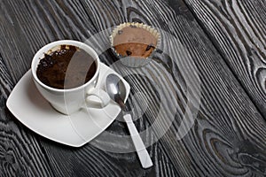 Coffee in a white cup stands on a saucer. Nearby lies a muffin, and a spoon on a saucer. On brushed pine boards
