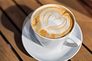 Coffee on wooden background. Cup of cappuccino with latte art on brown table with sunbeam. Fresh morning coffee with