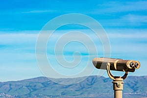 Coin operated binocular telescope for distant viewing. Blurred range of green hills, mountains in the distance under cloud sky