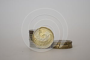 A coin worth twenty euro cents stands upright next to the other coins lying one on one on a white background