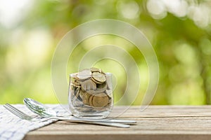 Coins in clear money jar, fork and spoon on wooden table with green blur light background. Savings money for eating concept