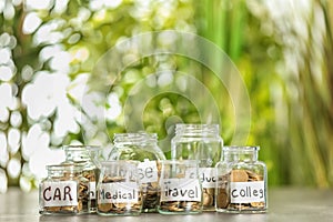 Coins for different needs in glass jars on table outdoors. Money savings concept