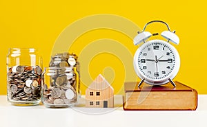 coins, small wooden house, white alarm clock and book placed on the white table Savings Ideas for Accounting and Finance
