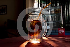 Coke inglass with straw. Brown sparkling beverage with sun backlight. Sparkling drink with ice in glass