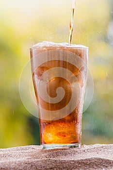 Cola water pouring into glass with ice cubes on sand