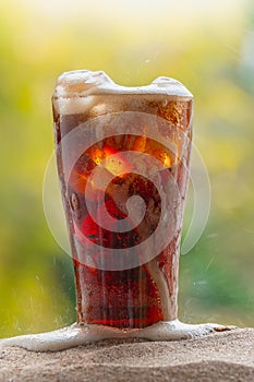 Cola water pouring into glass with ice cubes on sand