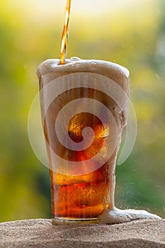 Cola water pouring into glass with ice cubes on sand