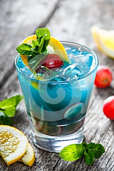 Cold blue alcoholic cocktail with lemon, grape and mint in glass on wooden background. Summer drinks