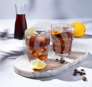 Cold brew coffee in a two glasses with lemon and ice on a marble board on a light background with coffee beans, bottle and shadows