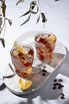 Cold brew coffee in a two glasses with lemon and ice on a marble board on a light background with coffee beans and shadows.