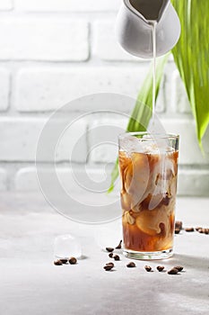 Cold brewed iced coffee in a tall glass cup and coffee beans on a gray concrete background. Pouring milk into a glass of coffee