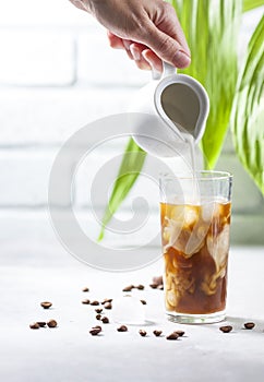 Cold brewed iced coffee in a tall glass cup and coffee beans on a gray concrete background. Pouring milk into a glass of coffee