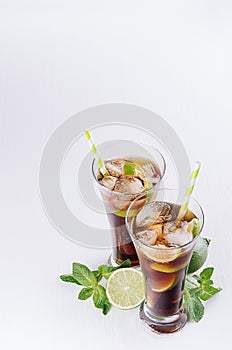 Cold cocktail cuba libre in two wet glasses with straw, pieces lime, mint on soft modern white background, top view, border.