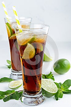 Cold cocktail cuba libre in two wet long glasses with straw, pieces lime, mint on soft modern white background.