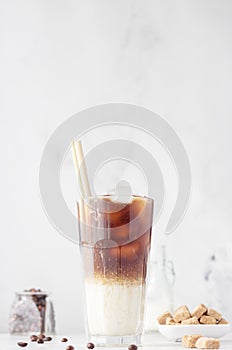 Cold coffee with milk and ice cubes in a tall glass with bamboo straw, light grey background.